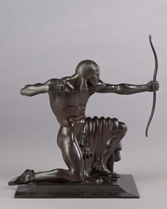 A bronze sculpture of a stylized Native American man on one knee pulling back the string of a bow he holds in front of him.