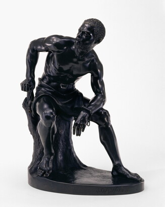 A bronze sculpture of a muscled Black man with manacles on his wrists leaning forward as he sits on a tree stump.