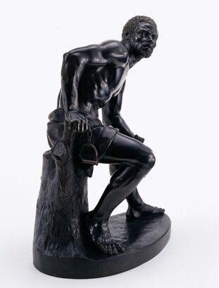 A bronze sculpture of a muscled Black man with manacles on his wrists leaning forward as he sits on a tree stump.