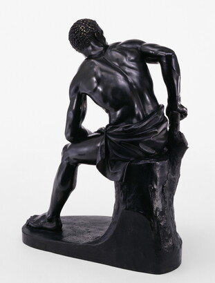 A bronze sculpture of a muscled Black man with manacles on his wrists leaning forward as he sits on a tree stump.