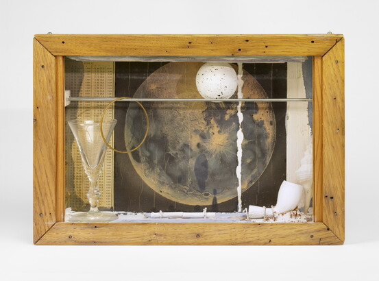 A wood shadowbox containing a pipe, a white ball with gray speckles, a picture of the moon, and a glass goblet.