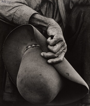 A close-up black-and-white photograph of a hand with aged, wrinkled skin holding a felt cowboy hat.