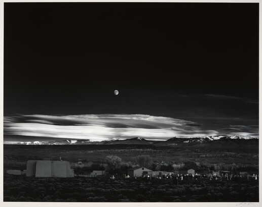 A black-and-white landscape photograph of a town with mountains in the background and the moon rising above the clouds in the night sky.