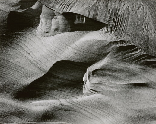An abstract black-and-white photograph of sand dunes with horizontal and vertical striations that make it seem to be in motion.