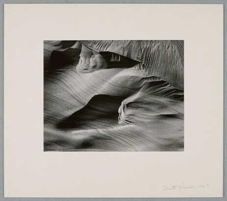 An abstract black-and-white photograph of sand dunes with horizontal and vertical striations that make it seem to be in motion.