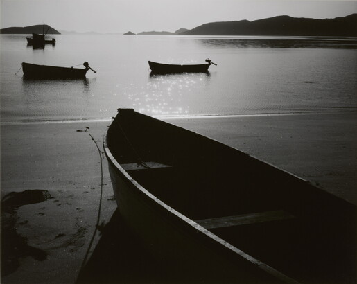 A black-and-white photograph of a rowboat on a sandy beach and three more out on still water with hills in the distance.