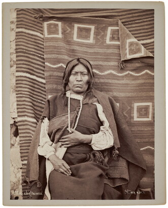 A sepia-toned photograph of an Indigenous person seated in front of woven blankets with their thumb hooked over a white beaded necklace hanging from their neck.