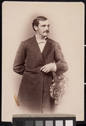 A sepia-toned portrait of a White man wearing a long frock coat standing posed with his arm on the back of a chair.