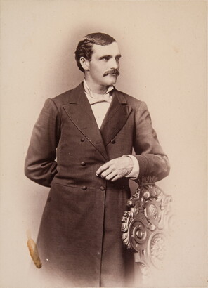 A sepia-toned portrait of a White man wearing a long frock coat standing posed with his arm on the back of a chair.