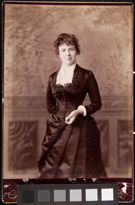 A black-and-white studio portrait photograph of a White woman in a formal, fitted dress standing in front of a backdrop.