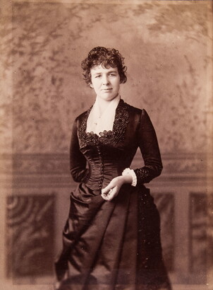 A black-and-white studio portrait photograph of a White woman in a formal, fitted dress standing in front of a backdrop.