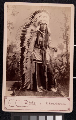 A sepia-toned studio portrait photograph of an Indigenous man in full traditional regalia, including a long, feathered headdress.