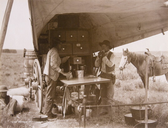 A sepia-toned photograph of two men eating and drinking from a chuckwagon as a horse stands in the background.