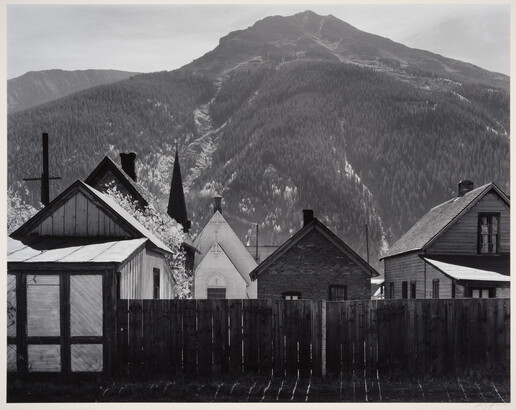 A black-and-white photograph of clapboard houses and a church behind a wood fence at the base of a densely wooded mountain.