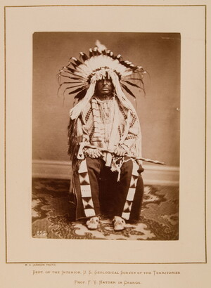 A black-and-white formal portrait photograph of a seated Indigenous man in full, traditional regalia with a headdress and tomahawk.