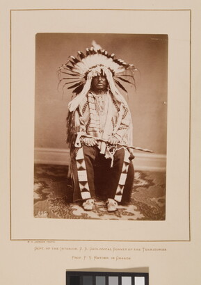 A black-and-white formal portrait photograph of a seated Indigenous man in full, traditional regalia with a headdress and tomahawk.