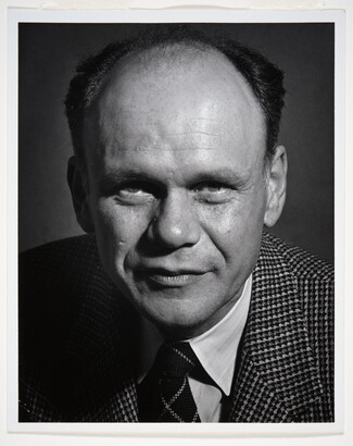 A black-and-white portrait photograph of a balding White man wearing a houndstooth blazer and striped tie.