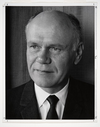 A black-and-white portrait photograph of a mostly bald White man gazing to the side wearing a dark jacket, dark tie, and white shirt.