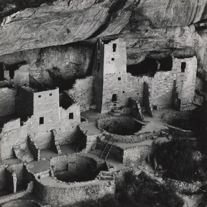 A black-and-white photograph of ruined stone buildings, some with towers, and several underground circular rooms all under the overhang of a cliff.