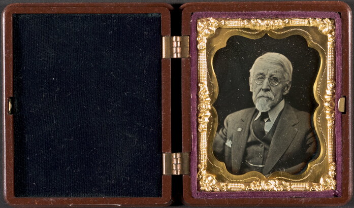 A black-and-white portrait photograph in a case with a decorative gold frame of an older, White man in a suit and glasses.