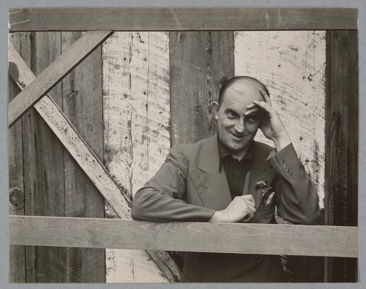 A black-and-white photograph of a bald, White man standing next to a wood structure, wearing a suit jacket and resting his head in his hand.
