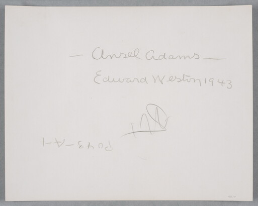 A black-and-white photograph of a bald, White man standing next to a wood structure, wearing a suit jacket and resting his head in his hand.