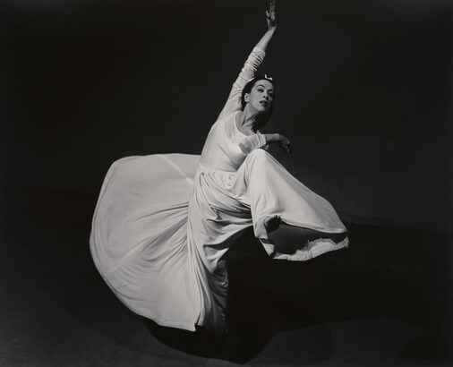 A black-and-white studio photograph of a woman wearing a long white dress that swirls as she dances.