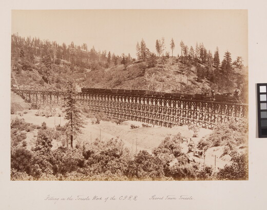 A sepia-toned photograph of a train travelling across a mountain valley with onlookers at base of the trestle.
