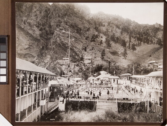 A black-and-white photograph of many people swimming in a large pool nestled at the base of a mountain.