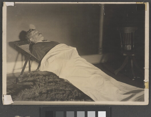 A black-and-white photograph of a White man in a formal suit laying on a stretcher and partially covered by a white sheet.