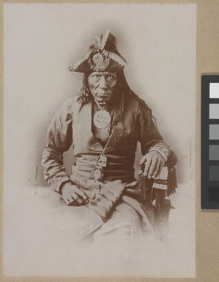 A black-and-white portrait photograph of a seated Indigenous man seated wearing a colonial-style jacket and a tricorn hat.