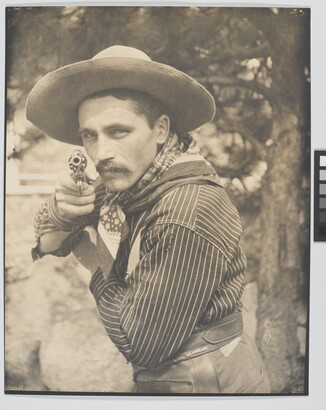 A sepia-toned photograph of a White man in a cowboy hat and bandana pointing a revolver at the camera.