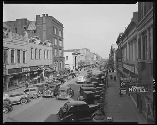 A black-and-white image of a street lined with tall buildings and parked automobiles.