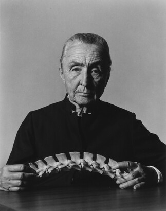 A black-and-white portrait photograph of an older White woman dressed in black seated at a table holding a skeletal spine.