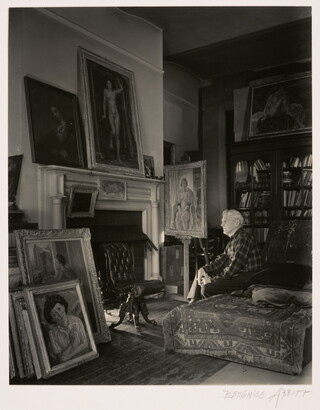 A black-and-white photograph of an older White man seated on a divan near a fireplace; elaborately framed paintings lean above the mantel, against the wall, and on an easel.