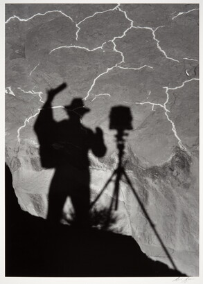 A black-and-white photograph of the shadow of a person and tripod with camera cast from above onto the landscape below.