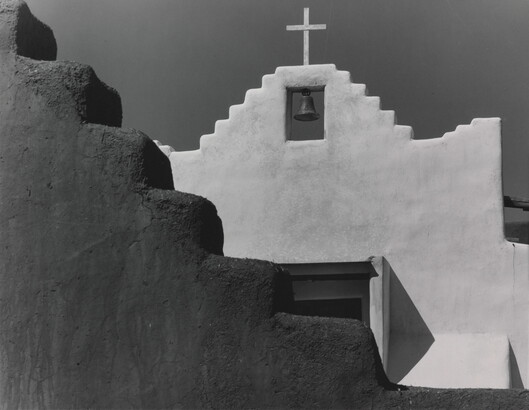 A black-and-white photograph of a terraced adobe bell tower with a cross on top, partially hidden by a terraced adobe wall.
