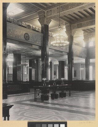 A black-and-white photograph of the interior of an ornate office lobby with inlaid floors, columns, and chandeliers.