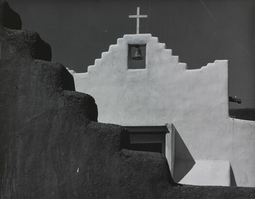 A black-and-white photograph of a terraced adobe bell tower with a cross on top, partially hidden by a terraced adobe wall.