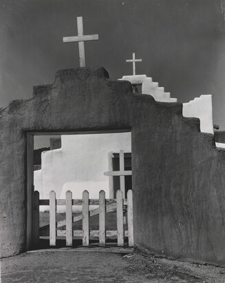 A black-and-white photograph of a white adobe church seen through the entrance of the surrounding adobe wall topped with a wooden cross.