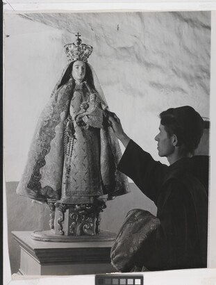 A black-and-white photograph of a friar touching a statue of the Virgin Mary dressed as queen of heaven holding the infant Jesus.