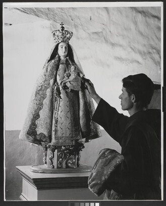 A black-and-white photograph of a friar touching a statue of the Virgin Mary dressed as queen of heaven holding the infant Jesus.