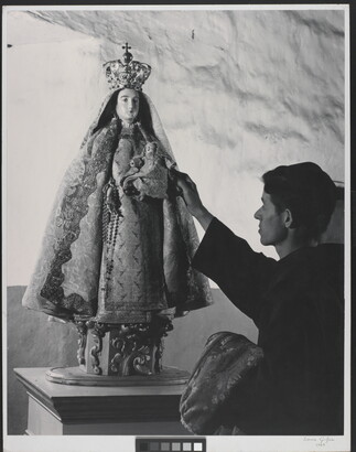 A black-and-white photograph of a friar touching a statue of the Virgin Mary dressed as queen of heaven holding the infant Jesus.
