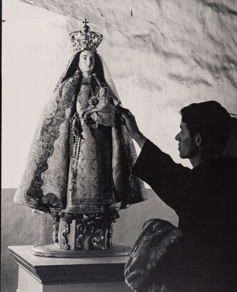A black-and-white photograph of a friar touching a statue of the Virgin Mary dressed as queen of heaven holding the infant Jesus.