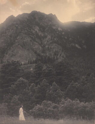 A black-and-white photograph of a woman in a white dress standing at the base of a rocky mountain covered with trees.