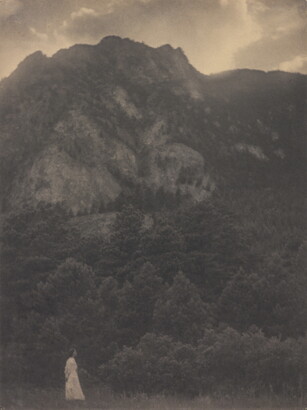 A black-and-white photograph of a woman in a white dress standing at the base of a rocky mountain covered with trees.