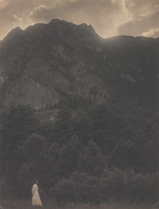 A black-and-white photograph of a woman in a white dress standing at the base of a rocky mountain covered with trees.