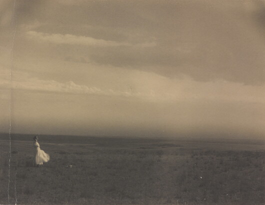 A black-and-white photograph of a woman in a white dress standing in a large meadow.