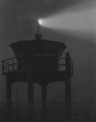 A black-and-white photograph of a person standing in silhouette atop a tower with a bright spotlight shining from the top.