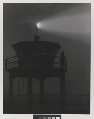 A black-and-white photograph of a person standing in silhouette atop a tower with a bright spotlight shining from the top.